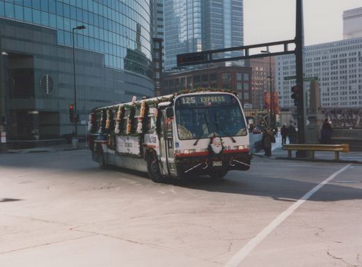 A white Chicago Transit Authority bus decorated with Christmas garlands and a Santa figure on the front, driving on a city street with modern buildings in the background.