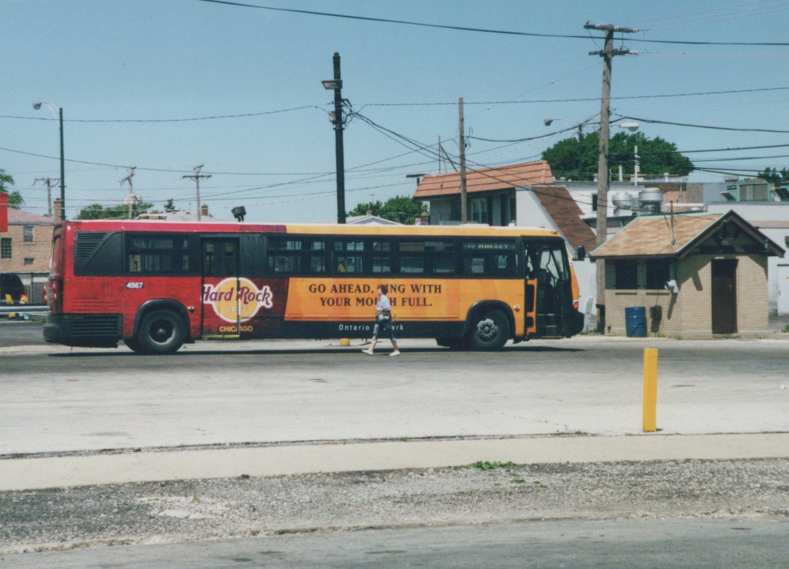 A red, orange, and black Chicago Transit Authority bus, number 4567, with a Hard Rock Cafe ad, is parked on a street with a pedestrian walking nearby on a sunny day.