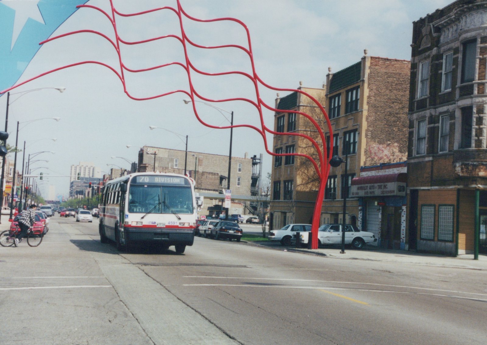 A white and red CTA bus drives down a city street under a large red, wavy sculpture, with buildings and parked cars along the sides.