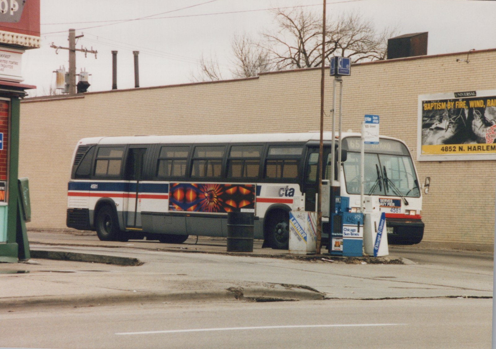 A white, blue and red CTA bus numbered 4581, with a '65 Grand' destination sign, is stopped at a street bus shelter next to newspaper boxes and a brick building.
