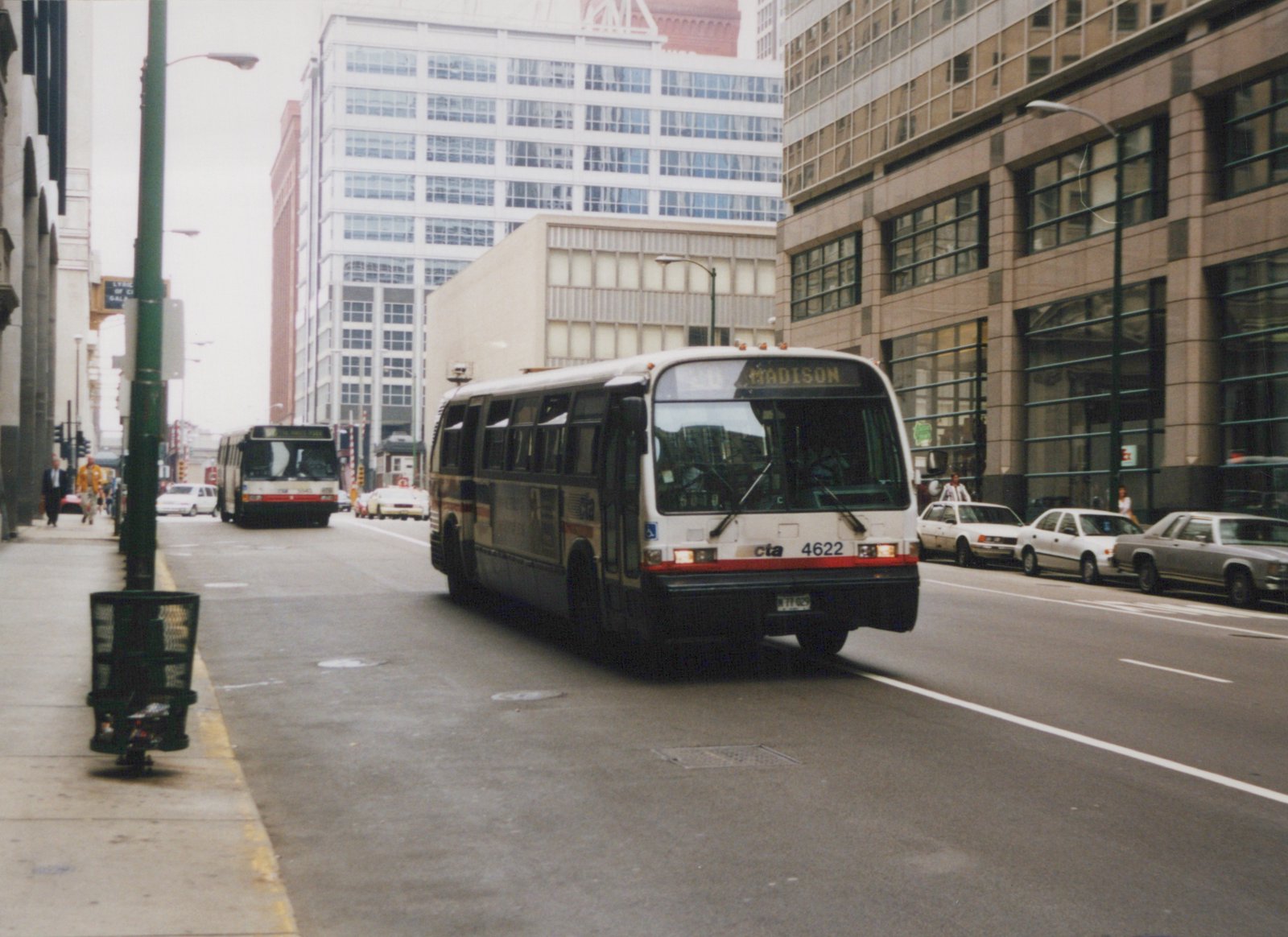 A white and red Chicago Transit Authority bus numbered 4622 is driving down a city street with buildings and other vehicles around it, in a cloudy day in 1997.