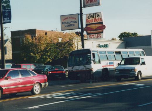 A white CTA bus numbered 4611 moves along a city street lined with cars and businesses, including a hot dog shop and a recruiting center, on a sunny autumn day.