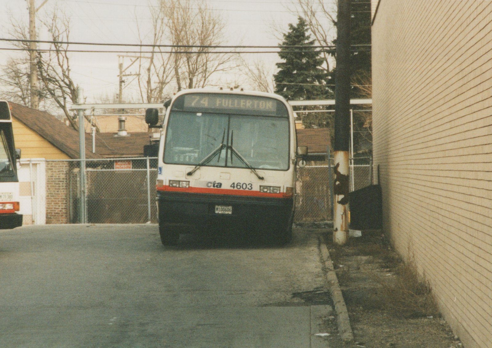 A white and red Chicago Transit Authority bus with the number 4603 on the front, parked on a street next to a brick wall, with bare trees and a chain-link fence in the background.
