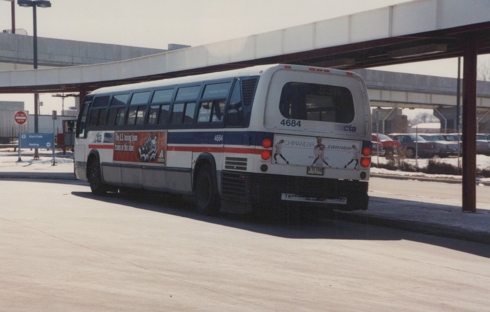 A white CTA bus numbered 4684, with blue and red stripes, is parked on a street under an elevated structure. Snow is visible on the ground, and the bus displays advertisements on its side and rear.