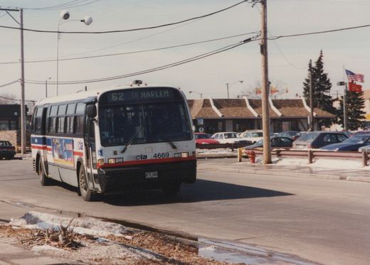 A white CTA bus with red and blue stripes is driving down a street on a clear winter day, with snow on the ground and cars parked in the background.