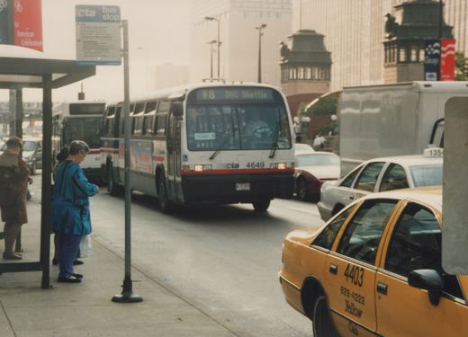 A white and red Chicago Transit Authority bus with the number 4649 is stopped on a city street next to a bus stop where people are waiting; yellow and white taxis are visible in the foreground and background.