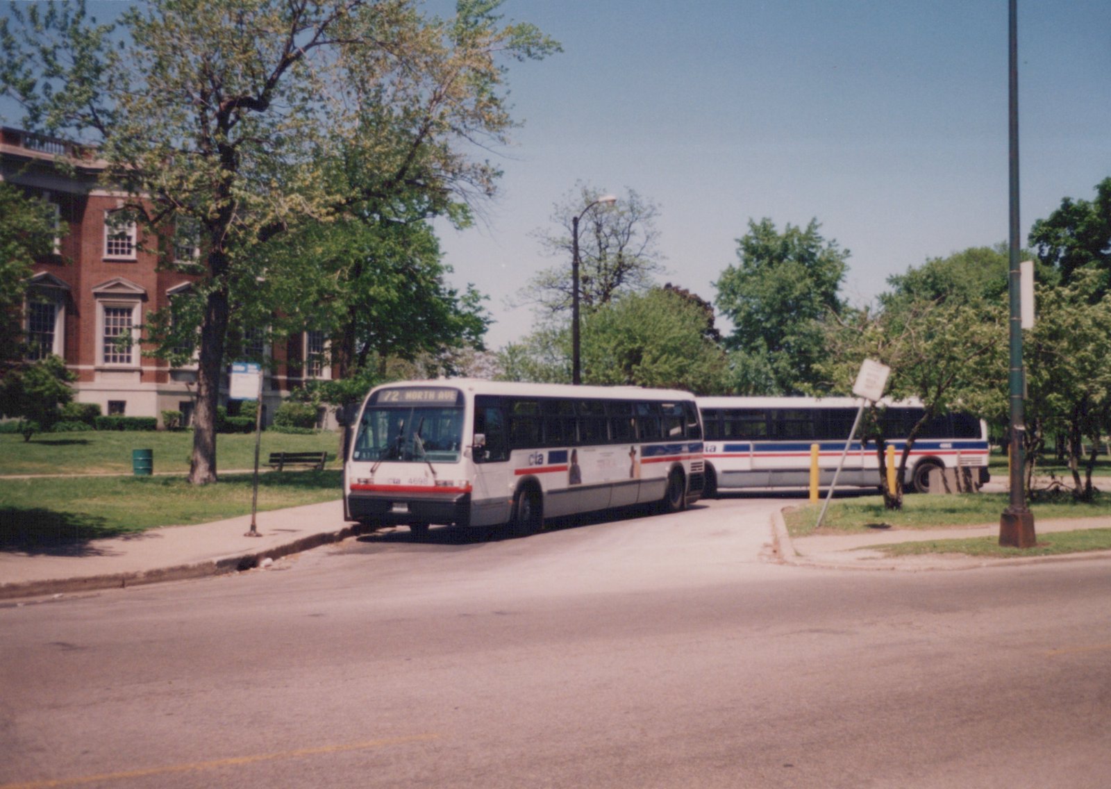 Two white Chicago Transit Authority buses with red and blue stripes on a city street, one showing route 72 North Avenue and fleet number 4698, parked near a park with a brick building in the background on a clear day.