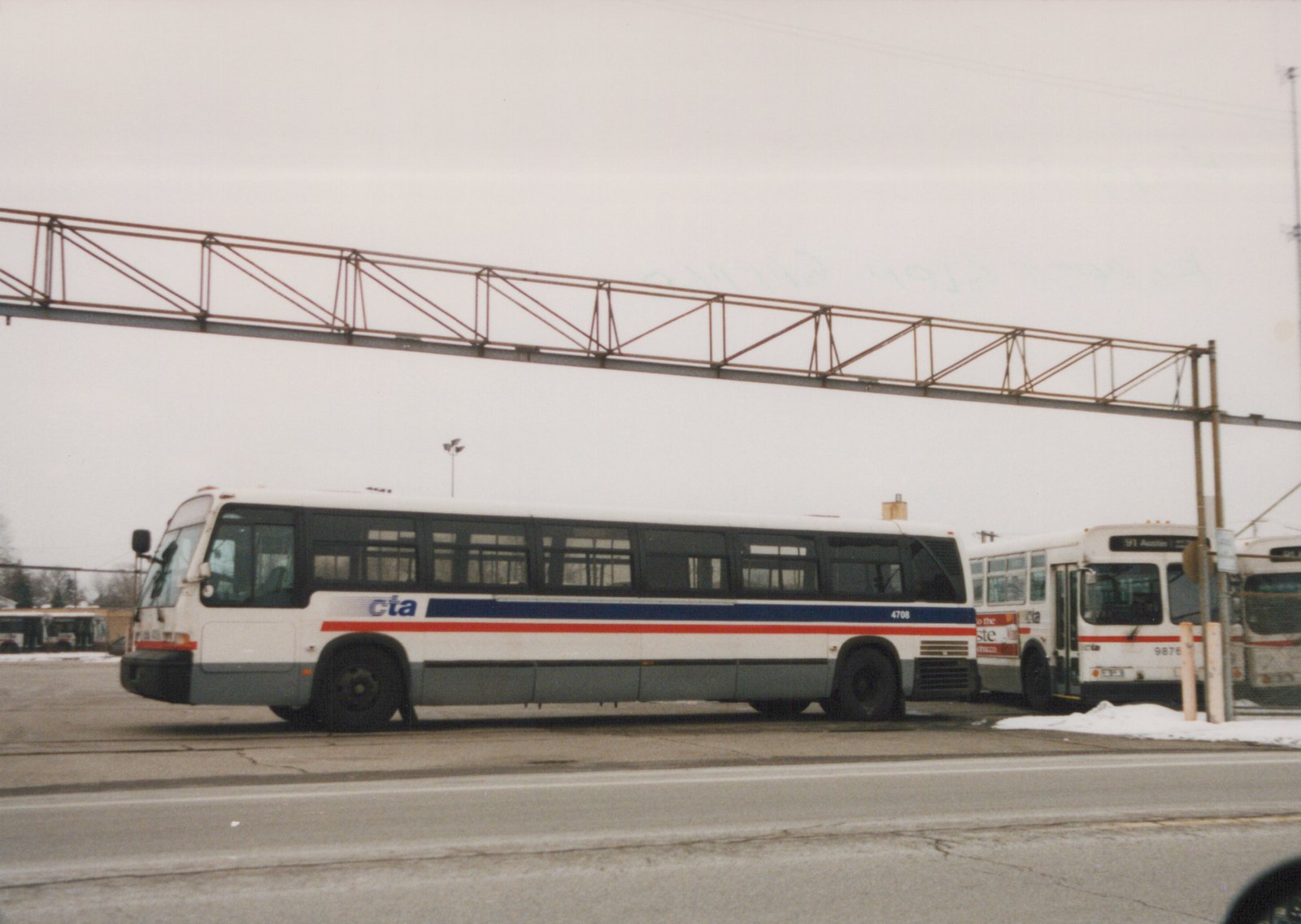 Side view of a white and gray CTA bus with blue and red stripes, number 4708, parked in a snowy lot with another CTA bus behind it and a large metal sign structure overhead.