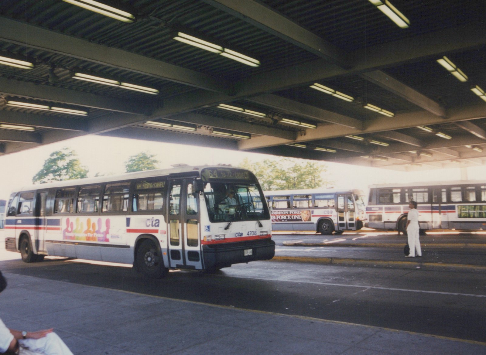 A white, blue, and red Chicago Transit Authority bus, number 4708, sits at a covered bus terminal with another bus visible behind it and a person standing on the pavement.