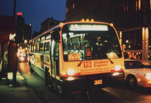 A white and blue CTA bus with number C84 and destination sign reading '19 Stadium Express' is shown on a city street at dusk, with its headlights on and a driver visible inside.