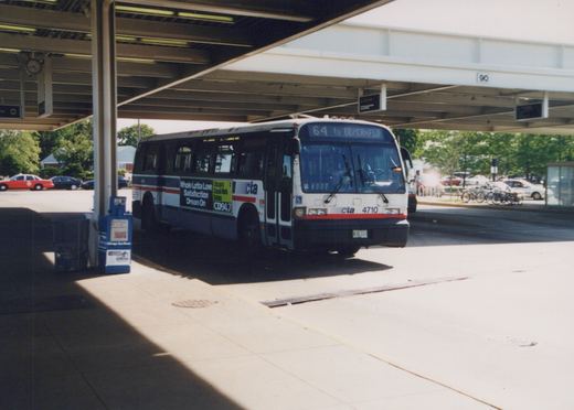 A white, blue, and red Chicago Transit Authority bus, number 4710, sits at a bus terminal under a covered structure on a sunny day, with a destination sign reading '64 to ER/CANFLD' and advertisements for CD94.7 on its side.