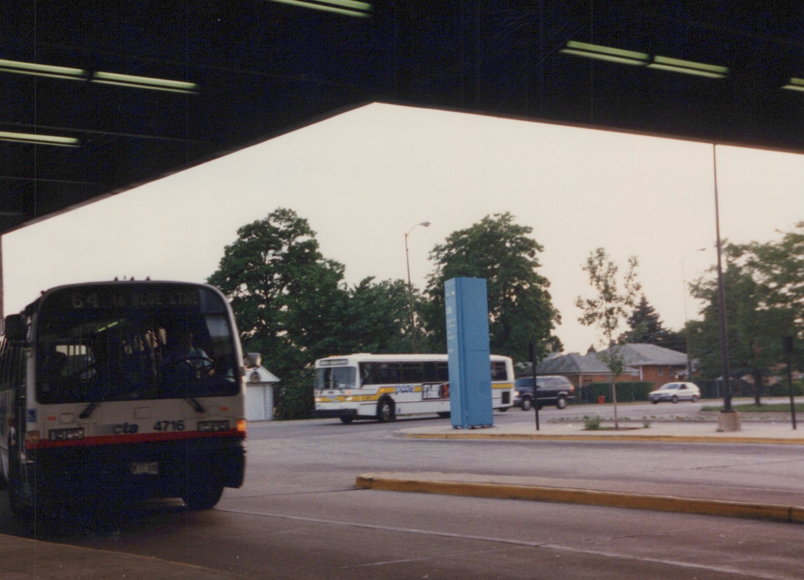 A white and red CTA bus, number 4716, is at a terminal. Another CTA bus is visible in the background along with a few cars, under an overcast sky.