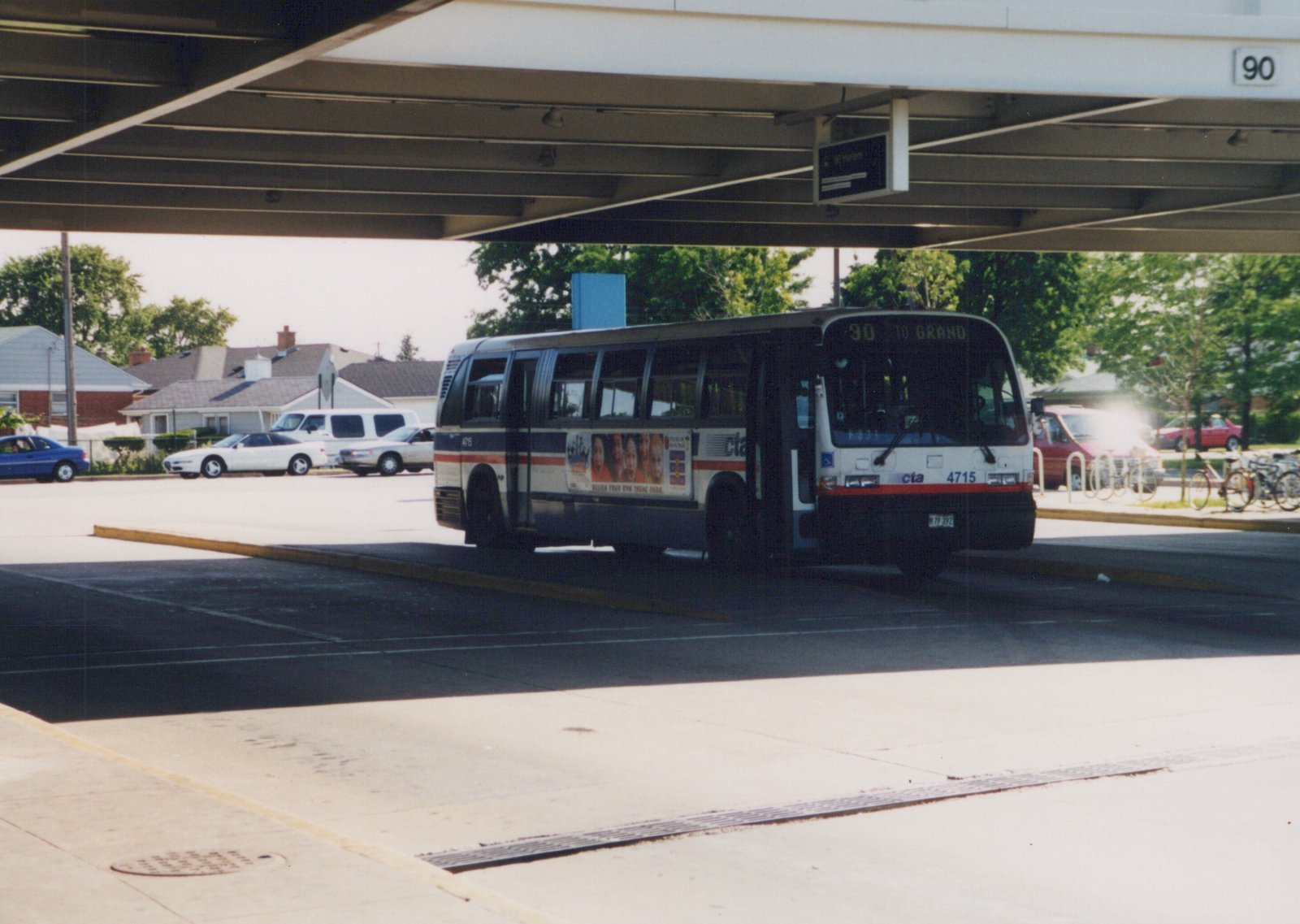 A white, blue, and red CTA bus numbered 4715 is parked at a bus terminal with the route '90 TO GRAND' displayed, under a large overhead structure with the number 90 on it, with cars and bicycles in the background.