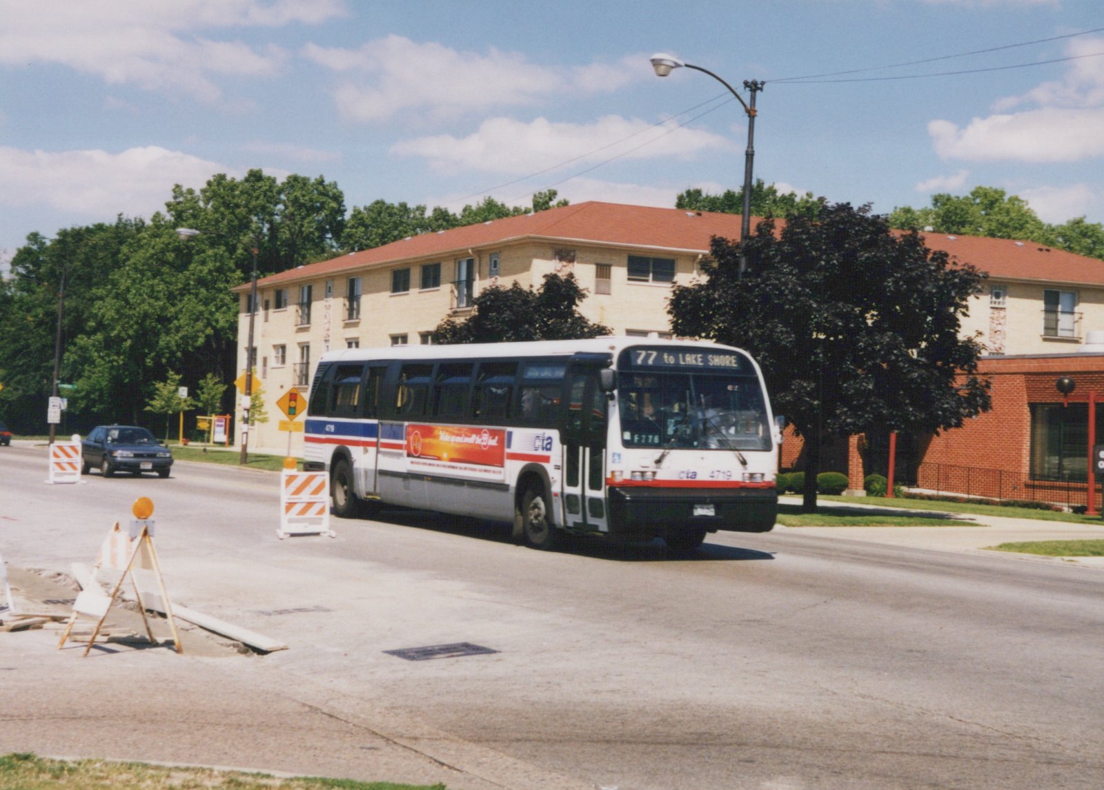 A white, red and blue Chicago Transit Authority bus, number 4719, drives down a street with a '77 to LAKE SHORE' sign on the front, passing construction barriers and a brick building under a blue sky.