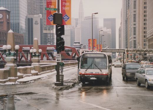 Front view of a white CTA bus with number 4719 on a wet, snowy city street with tall buildings and a red bridge in the background.