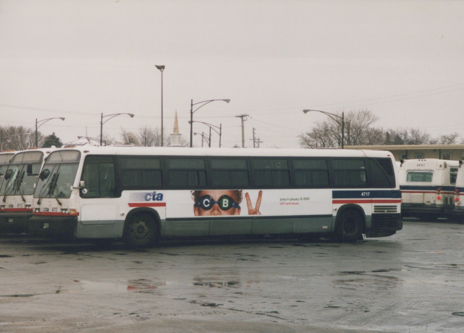 Side view of a white Chicago Transit Authority bus number 4717 parked in a wet lot, with a 'jump in january 15 2000' ad on its side, and other buses nearby under an overcast sky.