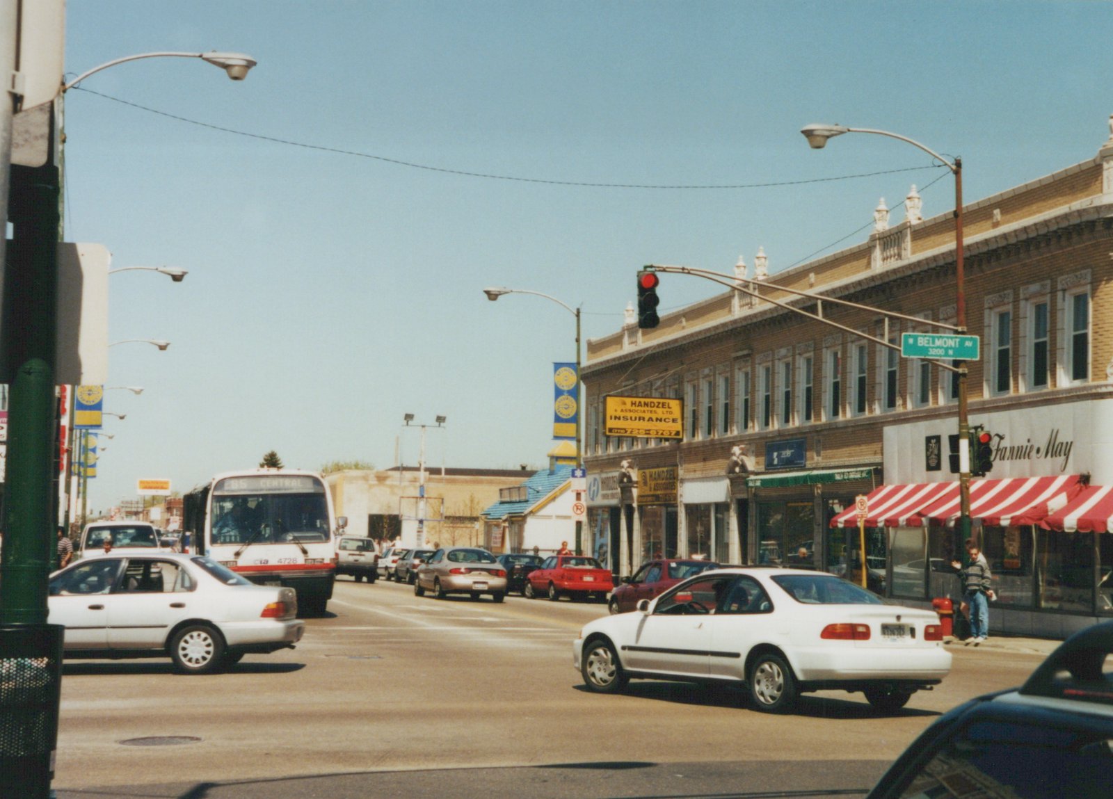 A white CTA bus with a red stripe, numbered 4728, drives down a city street lined with storefronts, including a Fannie May and an insurance agency, under a clear blue sky.