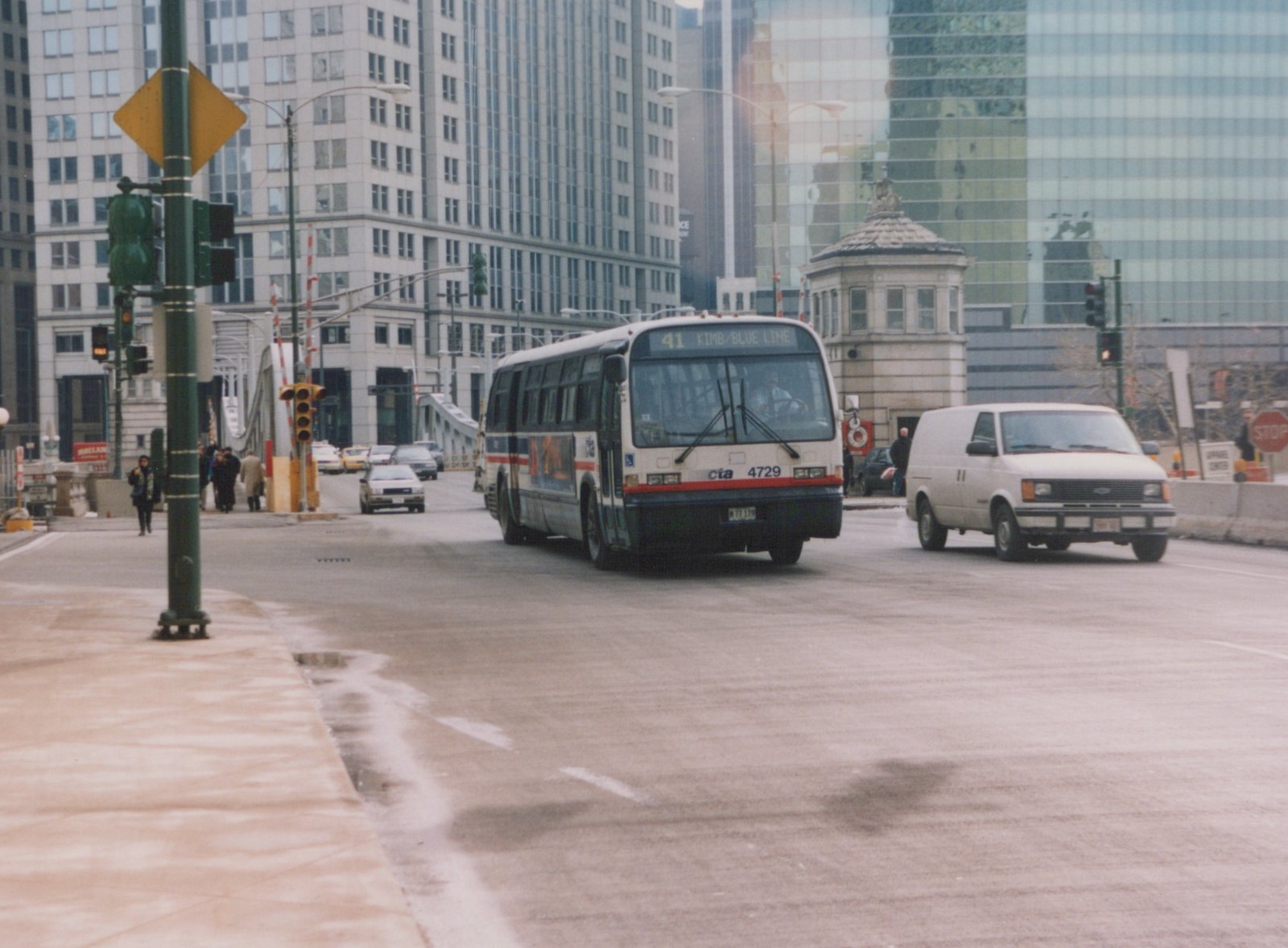 A white, blue, and red Chicago Transit Authority bus, number 4729, is driving on a city street next to a white van, with tall buildings and a bridge structure in the background.