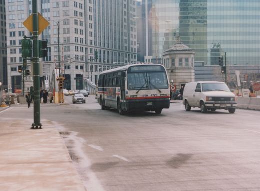 A white, blue, and red Chicago Transit Authority bus, number 4729, is driving on a city street next to a white van, with tall buildings and a bridge structure in the background.