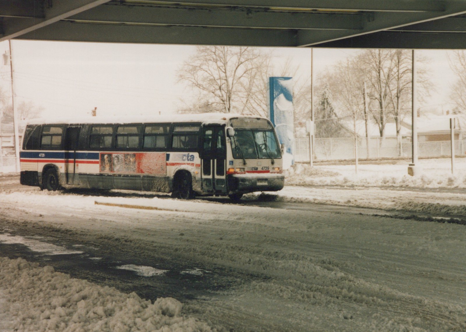 A white, red, and blue CTA bus numbered 4729 sits on a snow-covered street beneath a concrete overpass, with bare trees in the background.