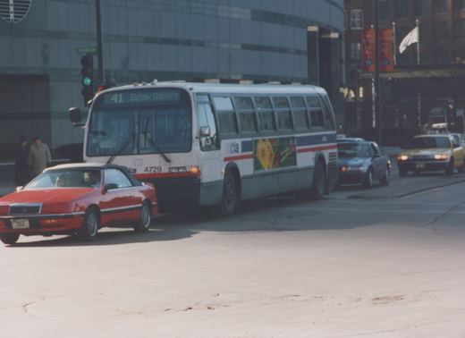 A white and blue Chicago Transit Authority bus, number 4729, on a city street with a red convertible and other cars, under a clear spring sky.