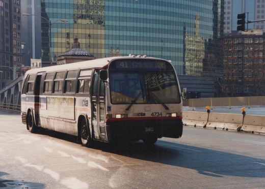A white and blue CTA bus numbered 4734 is driving on a city street with a modern glass building in the background.
