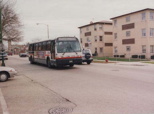 A white and blue Chicago Transit Authority bus, number 4736, is shown from the front three-quarters view driving down a street next to a tan brick apartment building on a cloudy day. Other cars are parked and driving nearby.