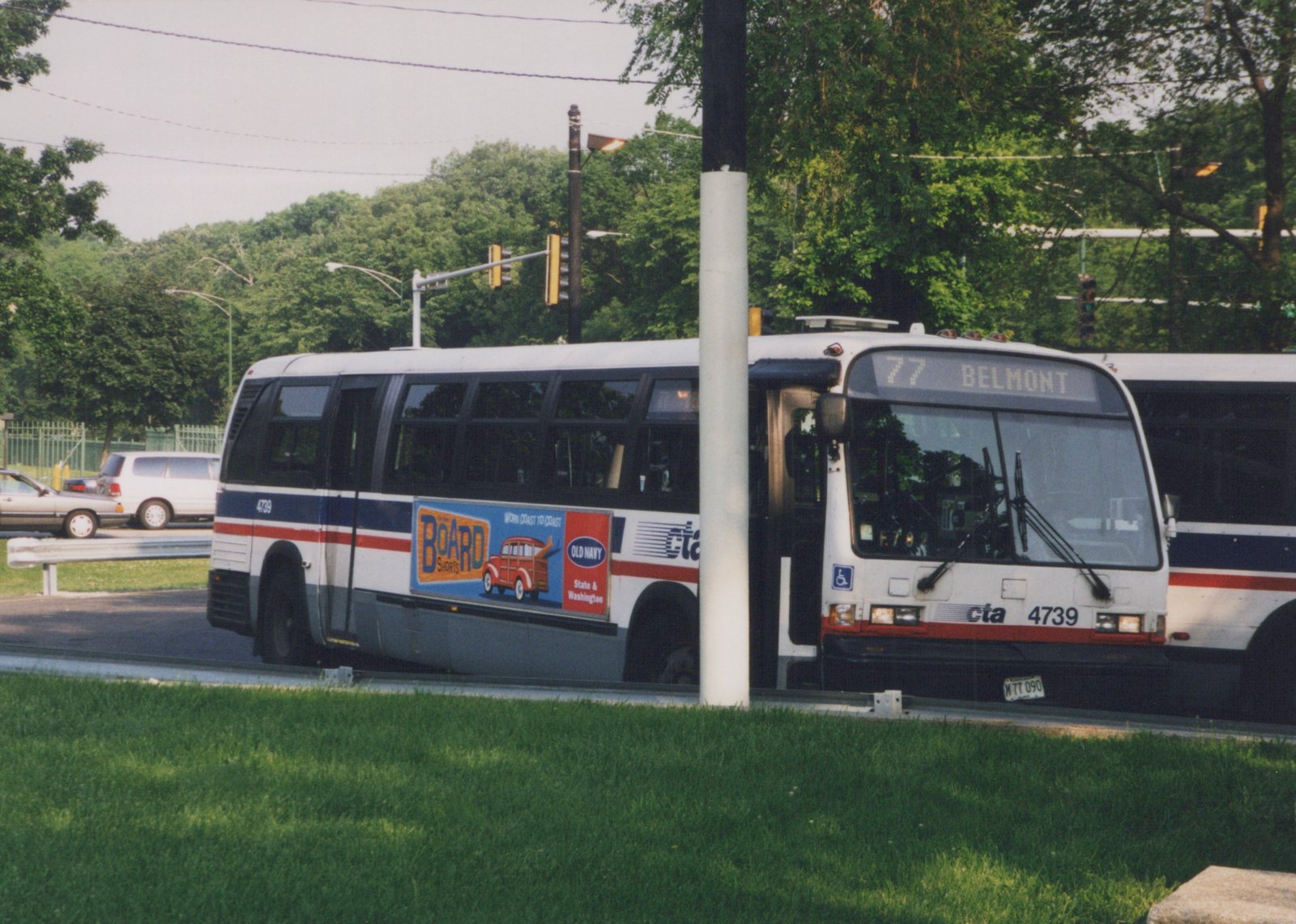 A side-front view of a white, blue, and red Chicago Transit Authority bus, number 4739, on a street with trees and a traffic light in the background, showing its destination as 77 Belmont.