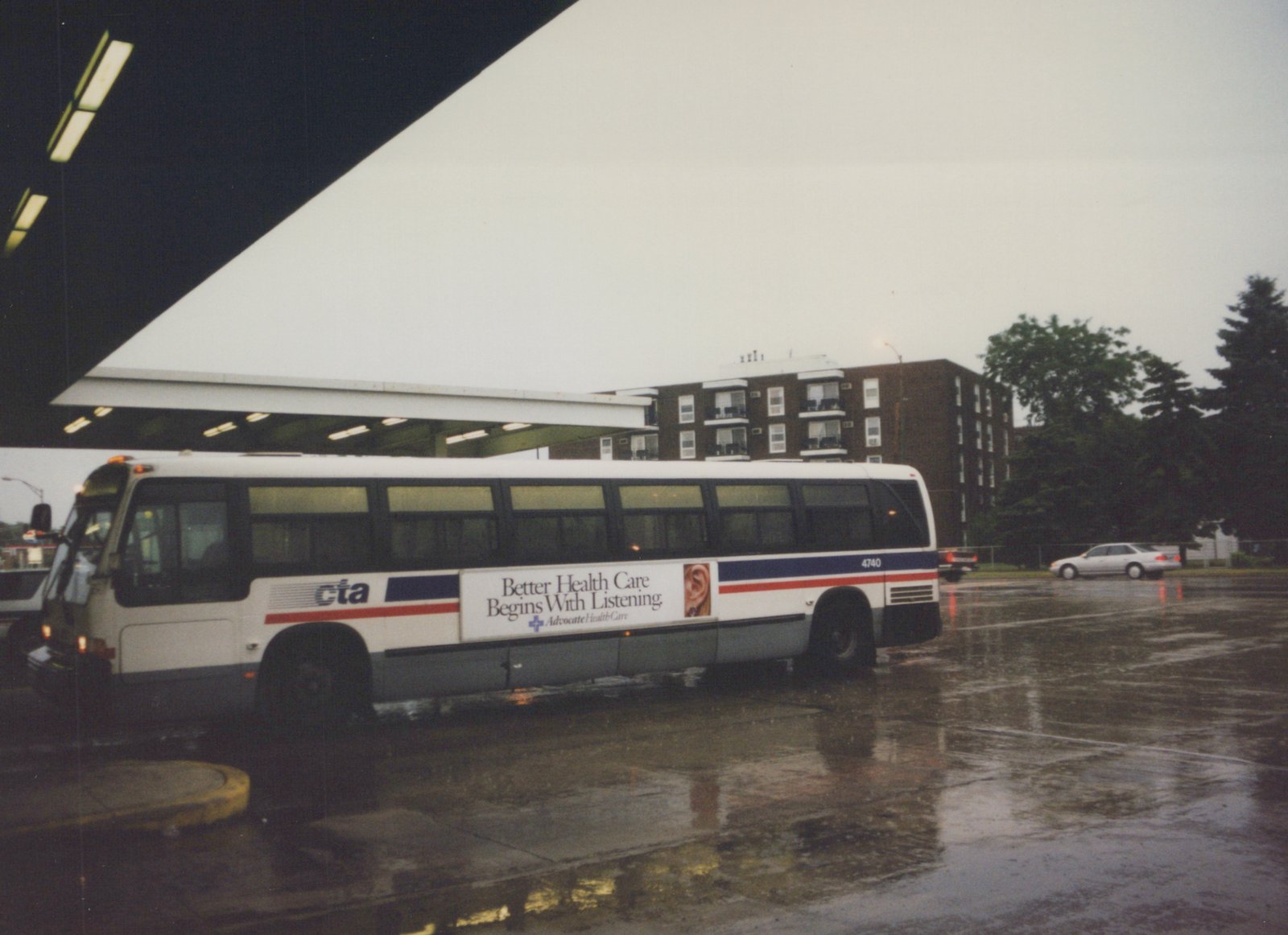 A side view of a white CTA bus with blue and red stripes, number 4740, parked on a wet pavement with a building and trees in the background.