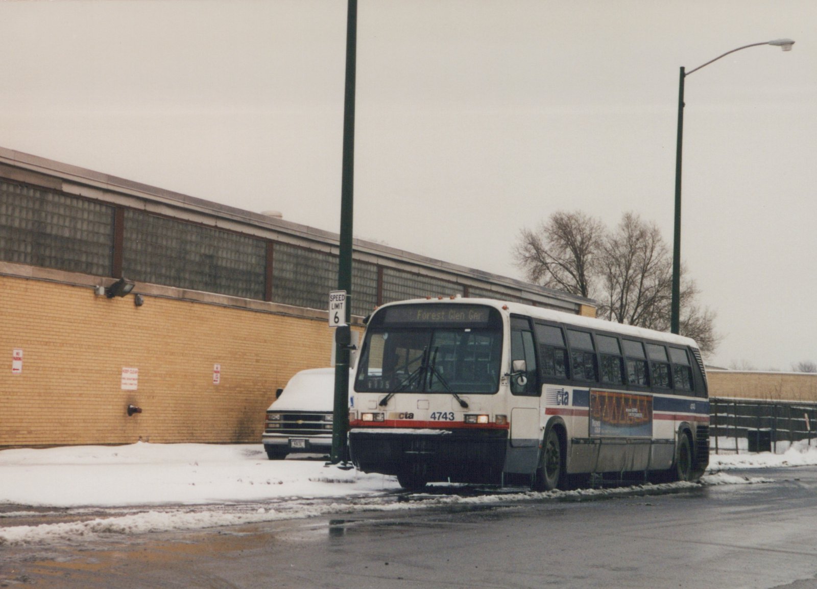 A white CTA motor bus, number 4743, is parked on a snowy street with a destination sign for Forest Glen Garage, in front of a large brick building.