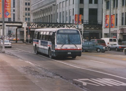 A white CTA bus with red and blue stripes, fleet number 4743, is on a city street under elevated train tracks, with other cars and pedestrians nearby on a cloudy winter day.