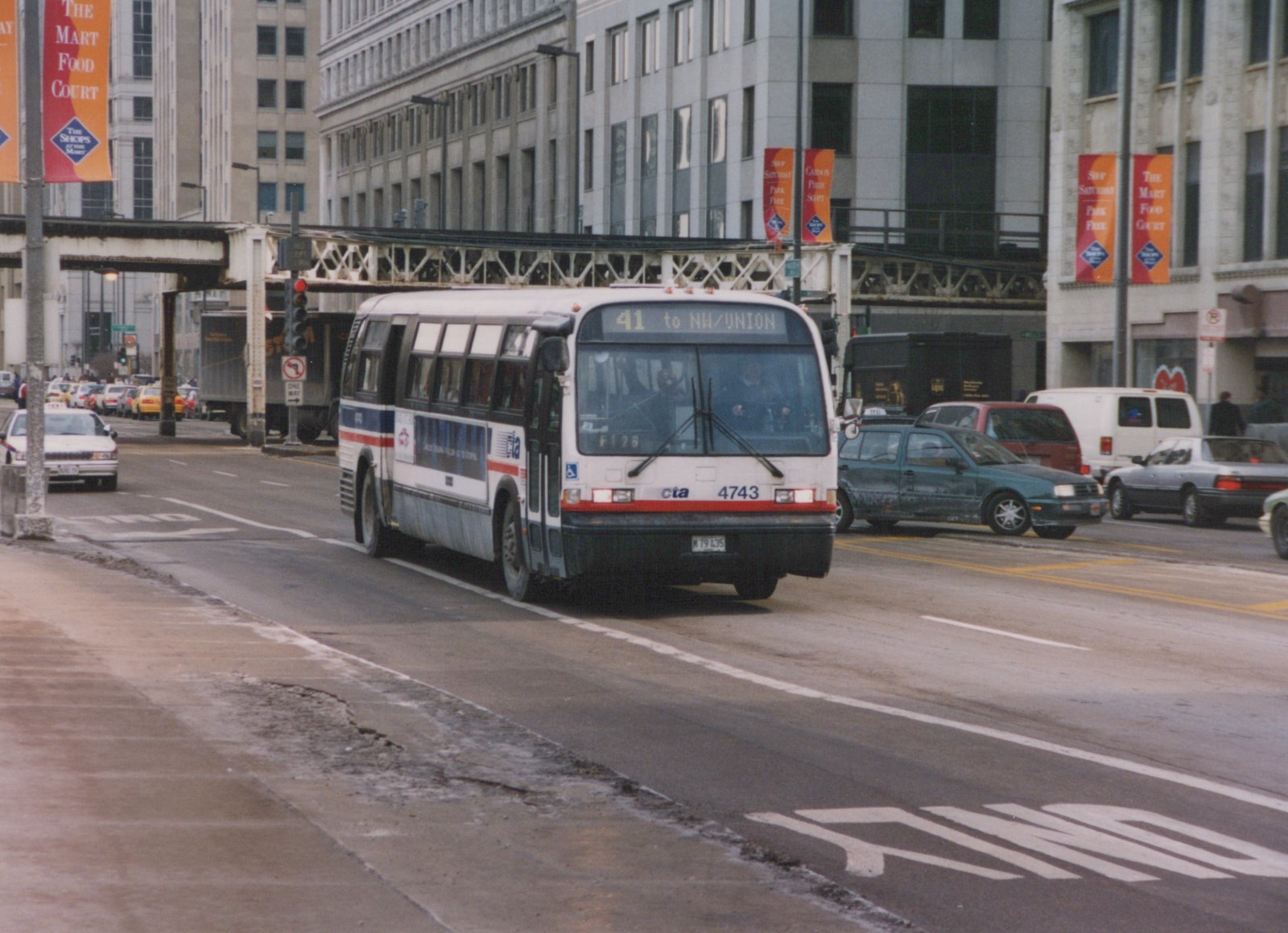 A white CTA bus with red and blue stripes, fleet number 4743, is on a city street under elevated train tracks, with other cars and pedestrians nearby on a cloudy winter day.
