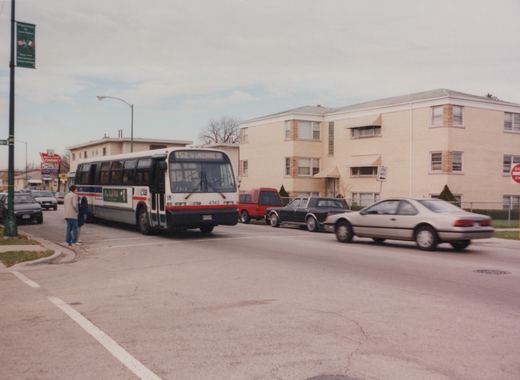 A white, blue and red Chicago Transit Authority bus, numbered 4743 and marked for route 152 to Lakeshore Drive, is stopped at a street corner with several cars and a two-story brick building in the background.