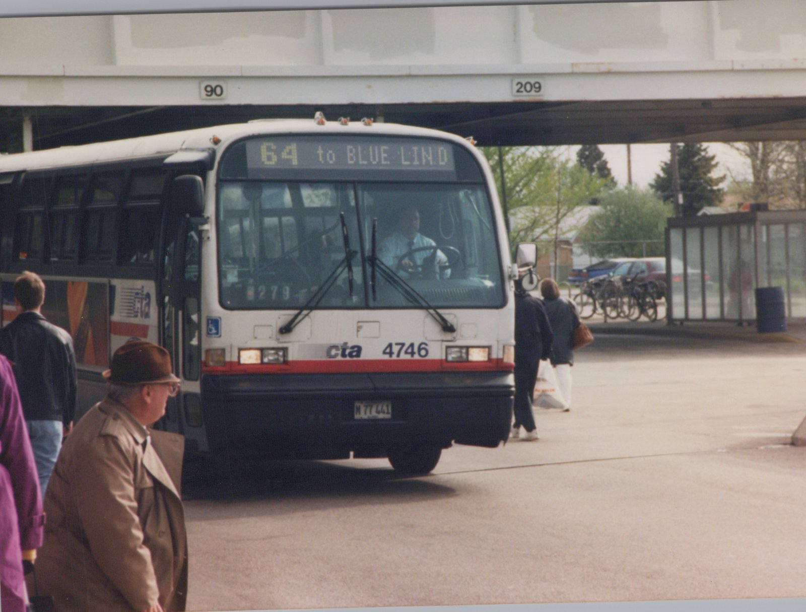 A white and red CTA bus with the number 4746 and route 64 to Blue Island on its sign is stopped under an overpass as passengers board. People are walking nearby, and bicycles are parked in the background.