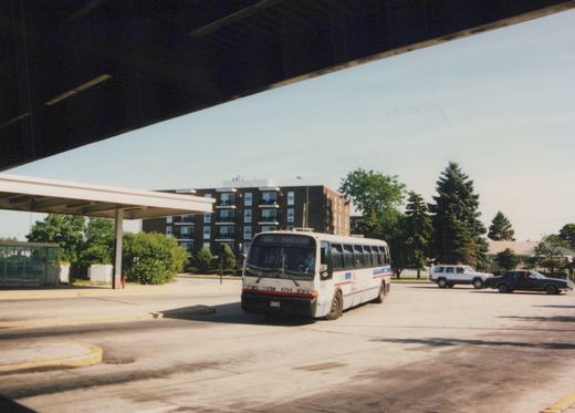 A white Chicago Transit Authority bus with red and blue stripes, number 4744, is shown from a three-quarter front view under a concrete overpass on a sunny day, with apartment buildings in the background.