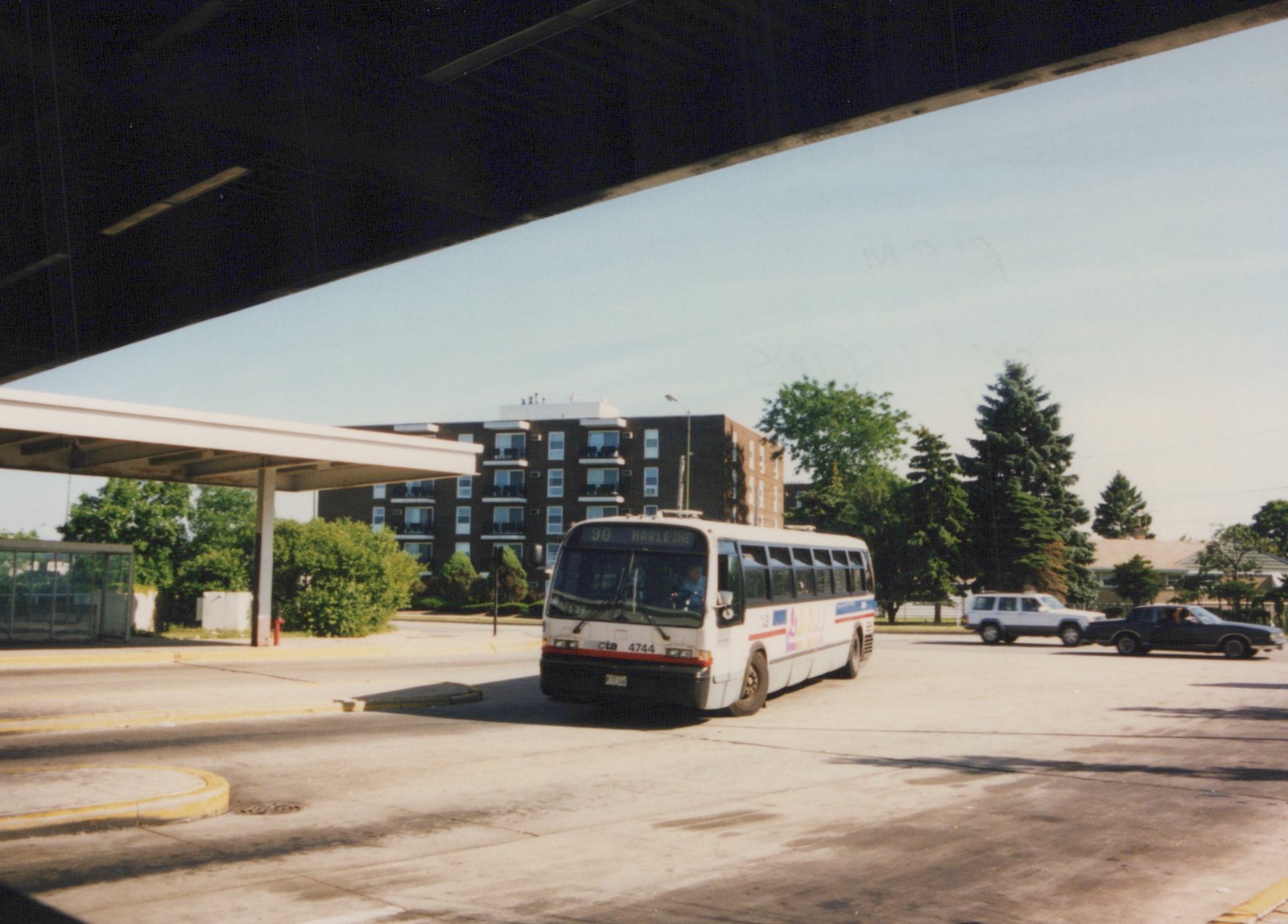 A white Chicago Transit Authority bus with red and blue stripes, number 4744, is shown from a three-quarter front view under a concrete overpass on a sunny day, with apartment buildings in the background.