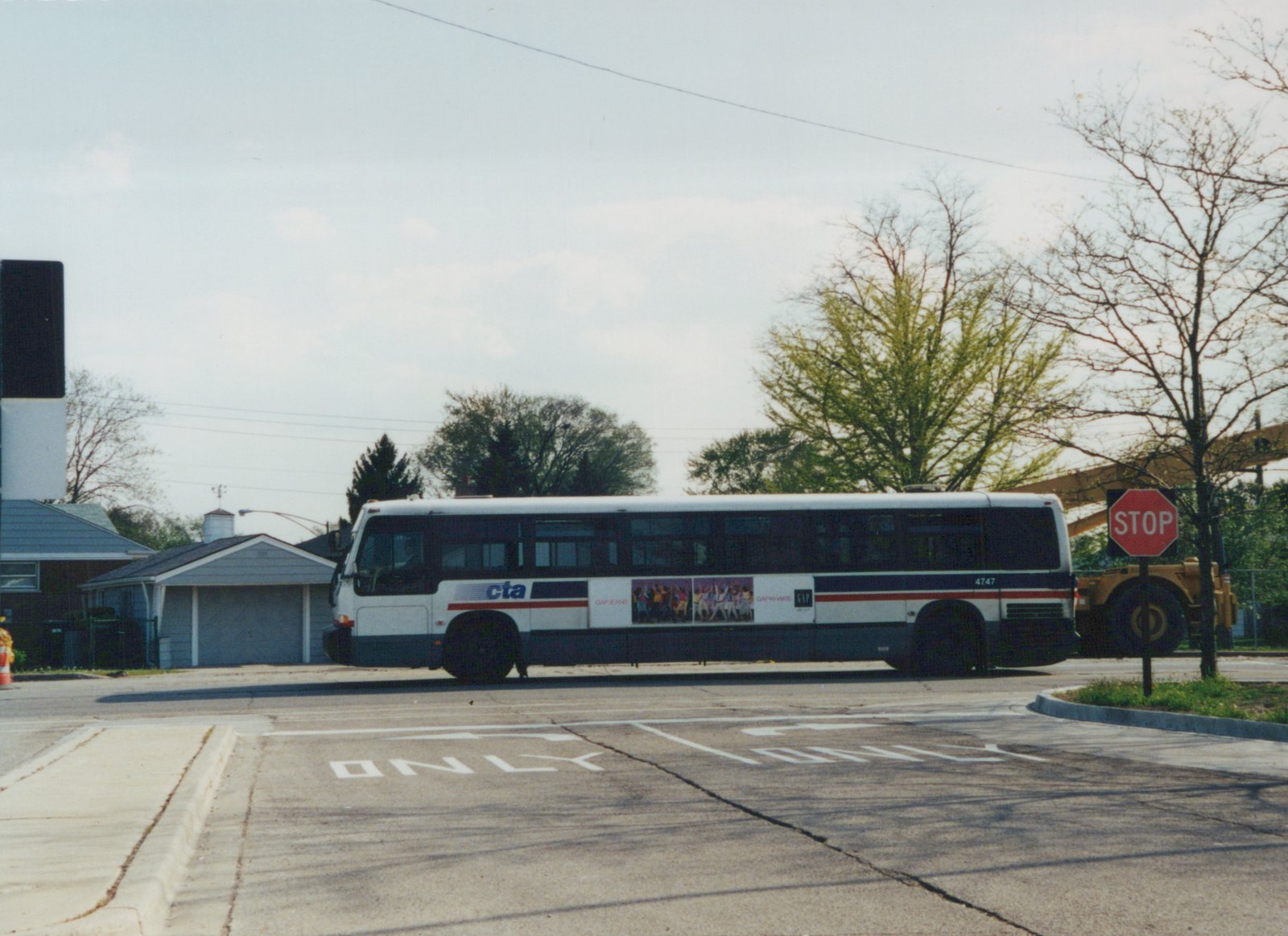 A white, blue, and red Chicago Transit Authority bus numbered 4747 is parked on a street corner next to a house and a stop sign, with trees in early spring foliage behind it.