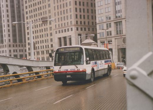 A white and blue Chicago Transit Authority bus, number 4749, is driving on a wet bridge with tall buildings in the background.