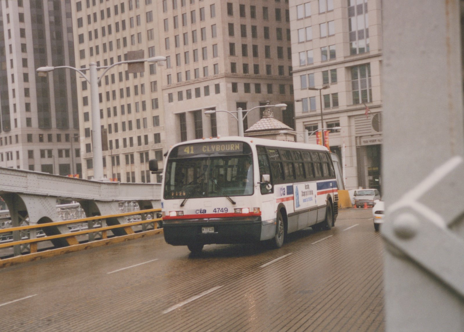 A white and blue Chicago Transit Authority bus, number 4749, is driving on a wet bridge with tall buildings in the background.