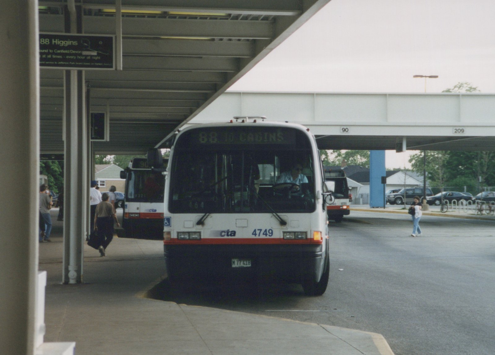 A white and red CTA bus with the number 4749 is parked at a bus terminal under an overpass, with people walking on the platform and another bus visible behind it.
