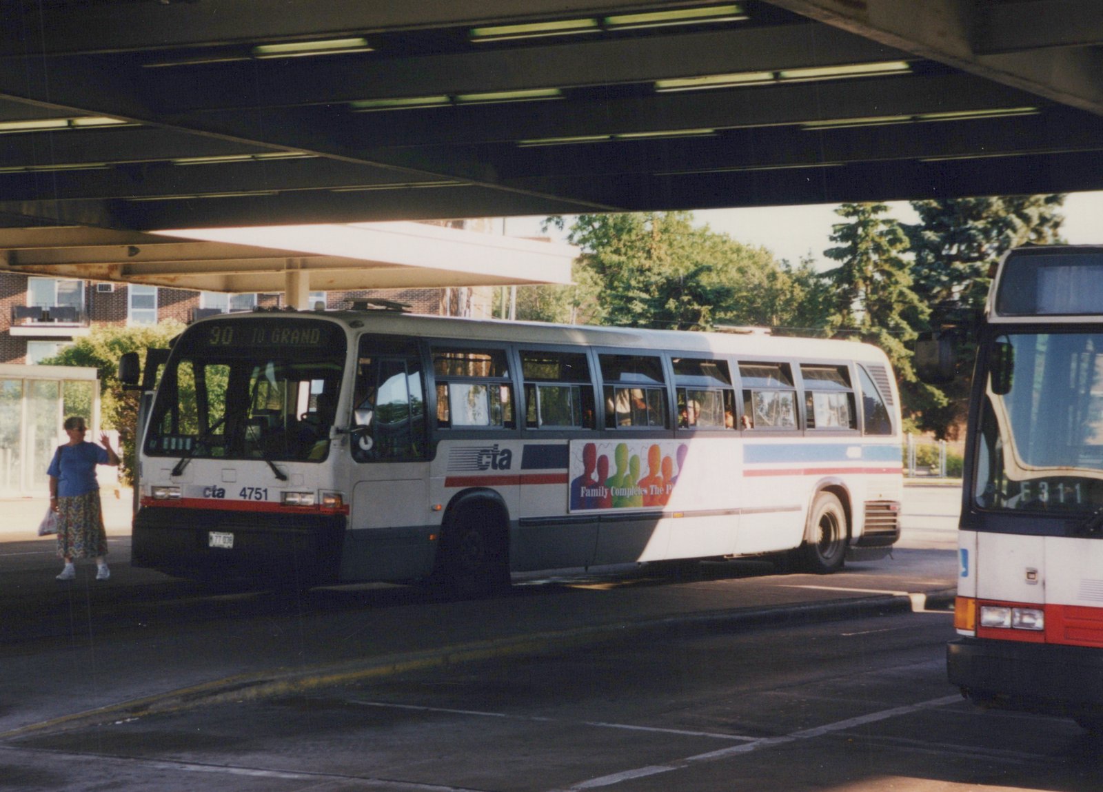 A white and blue CTA bus with the number 4751 is parked under a concrete overpass on a sunny day, with a woman standing nearby and another bus partially visible to the right.