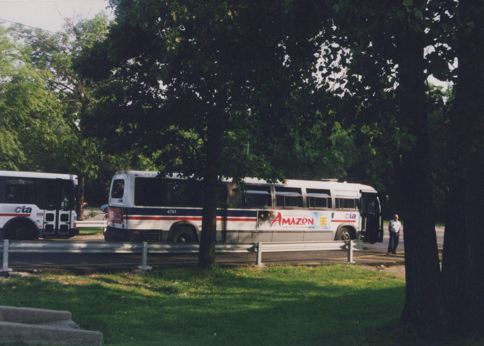 A white CTA bus with blue and red stripes, number 4751, is parked on a street with green trees and grass, a man standing near its door.