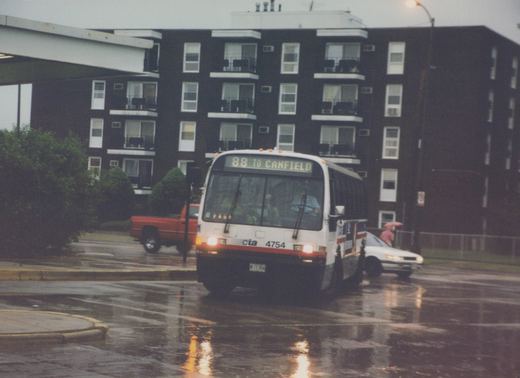 A white CTA bus with red and blue stripes, numbered 4754 and displaying route 88 to Canfield, drives down a wet urban street with a dark apartment building in the background. A person with a red umbrella stands nearby, and a red pickup truck is visible behind the bus.