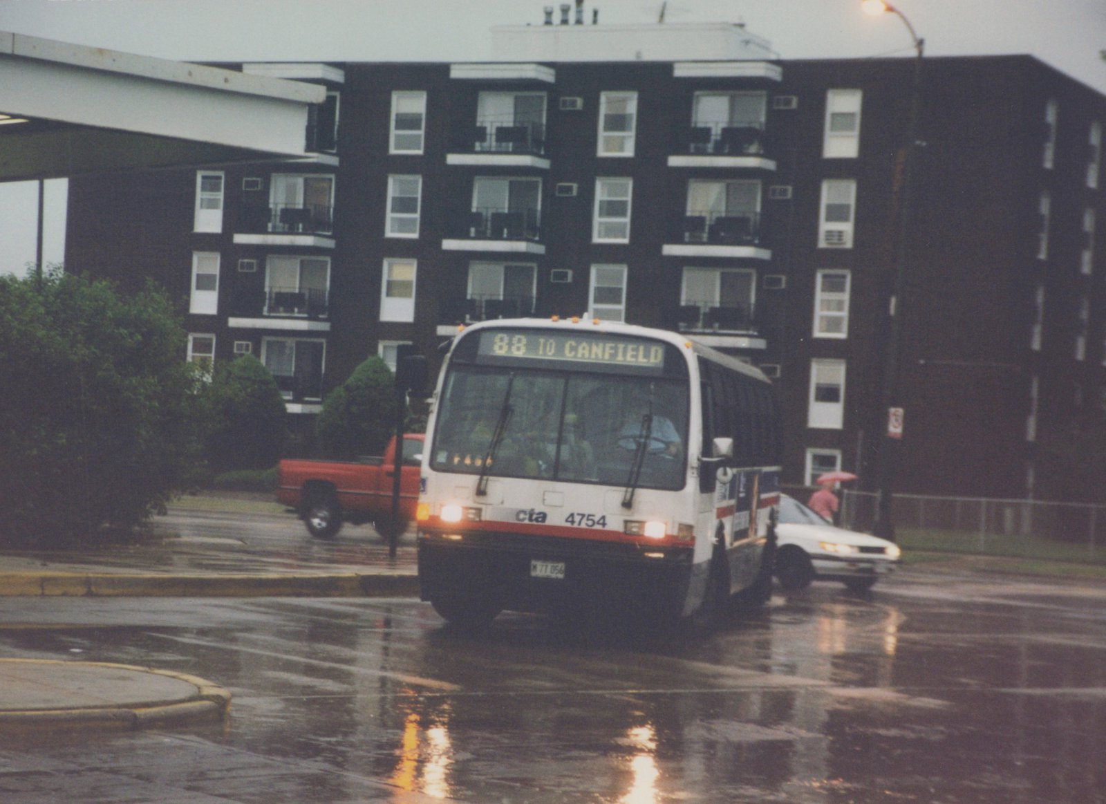 A white CTA bus with red and blue stripes, numbered 4754 and displaying route 88 to Canfield, drives down a wet urban street with a dark apartment building in the background. A person with a red umbrella stands nearby, and a red pickup truck is visible behind the bus.