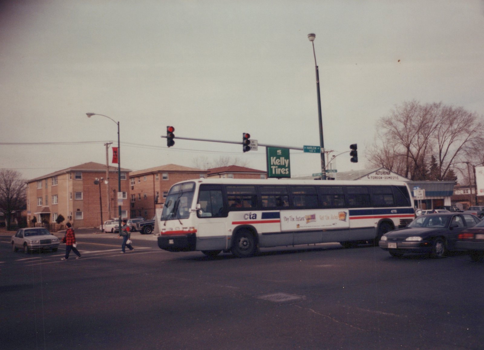 A white, blue, and red CTA bus numbered 4779 is turning at a city street intersection with pedestrians crossing and bare trees in the background.