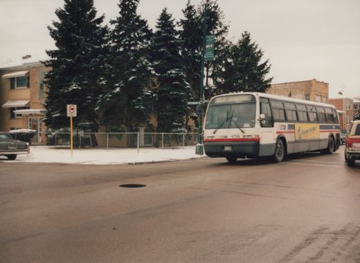 A white CTA bus with red and blue stripes and the number 4799 turns at an intersection with snow on the ground and trees, with other cars nearby.