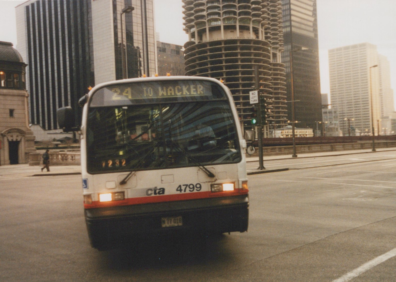 A white and red CTA bus numbered 4799 is driving on a city street with tall buildings, including Marina City, in the background. The destination sign reads '24 TO WACKER'.