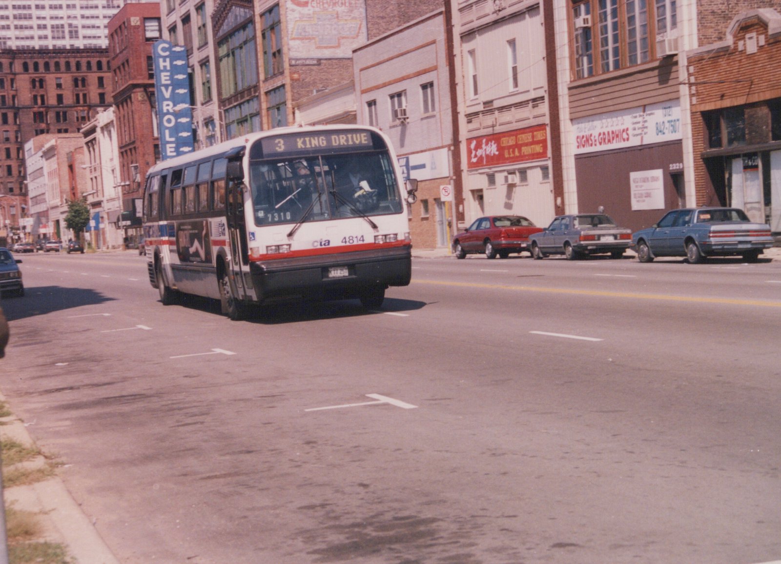 A white, red, and blue Chicago Transit Authority bus with the number 4814 is driving down a city street with parked cars and storefronts in the background.