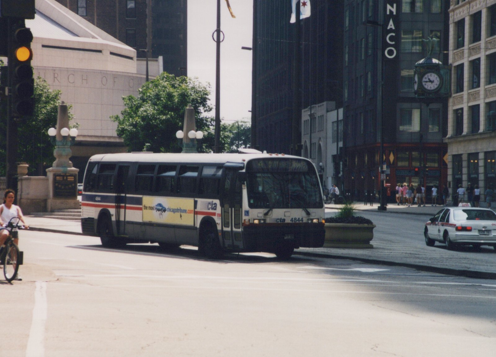 A white and blue CTA bus with the number 4844 and a 'Cottage Grove' destination sign is driving down a city street, with a taxi and a cyclist nearby, and tall buildings in the background.