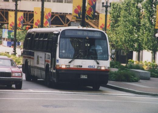A white CTA bus with the number 4862 on the front and a 'NOT IN SERVICE' sign above the windshield is driving on a city street next to a maroon car, with green trees and yellow banners in the background.
